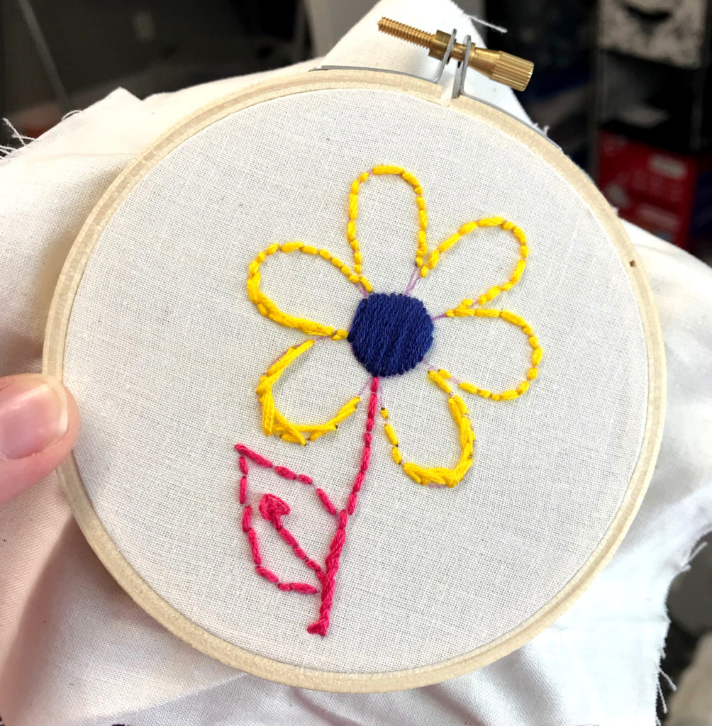Picture of a small hoop with a brightly colored flower stitched on it. 