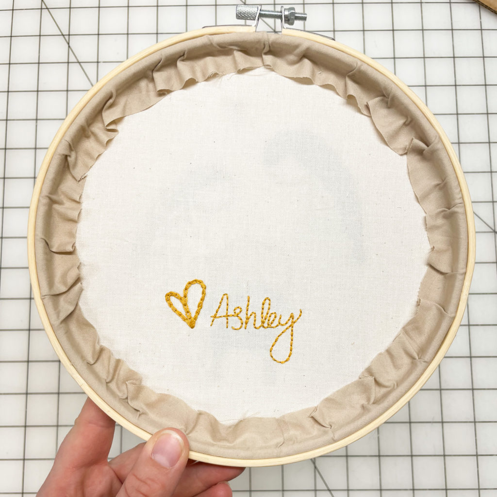 Finishing back of embroidery hoop with stitching that says "heart, Ashley."