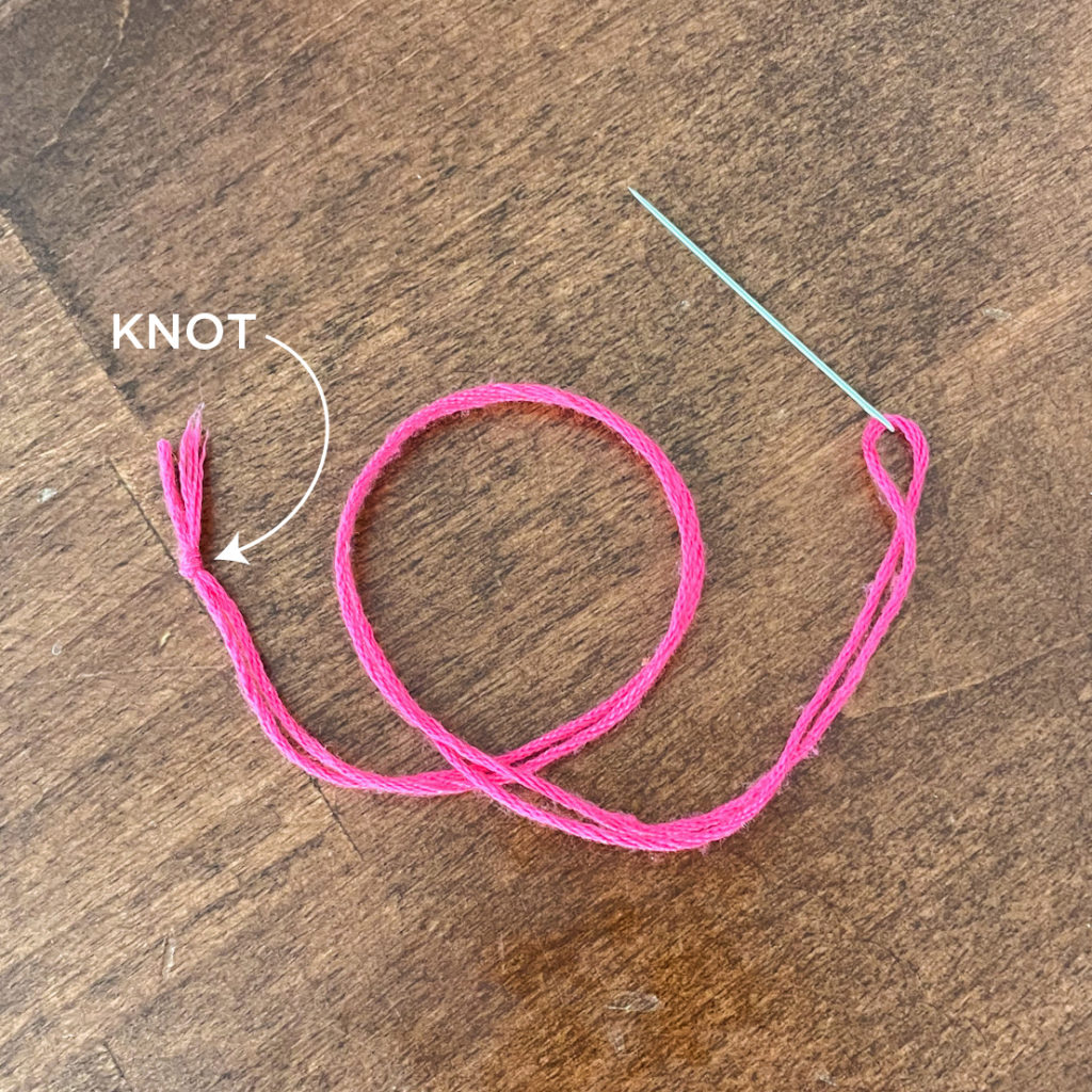 Image shows pink floss threaded through a needle. Both ends of the thread are tied together. An arrow points to the knot. 
