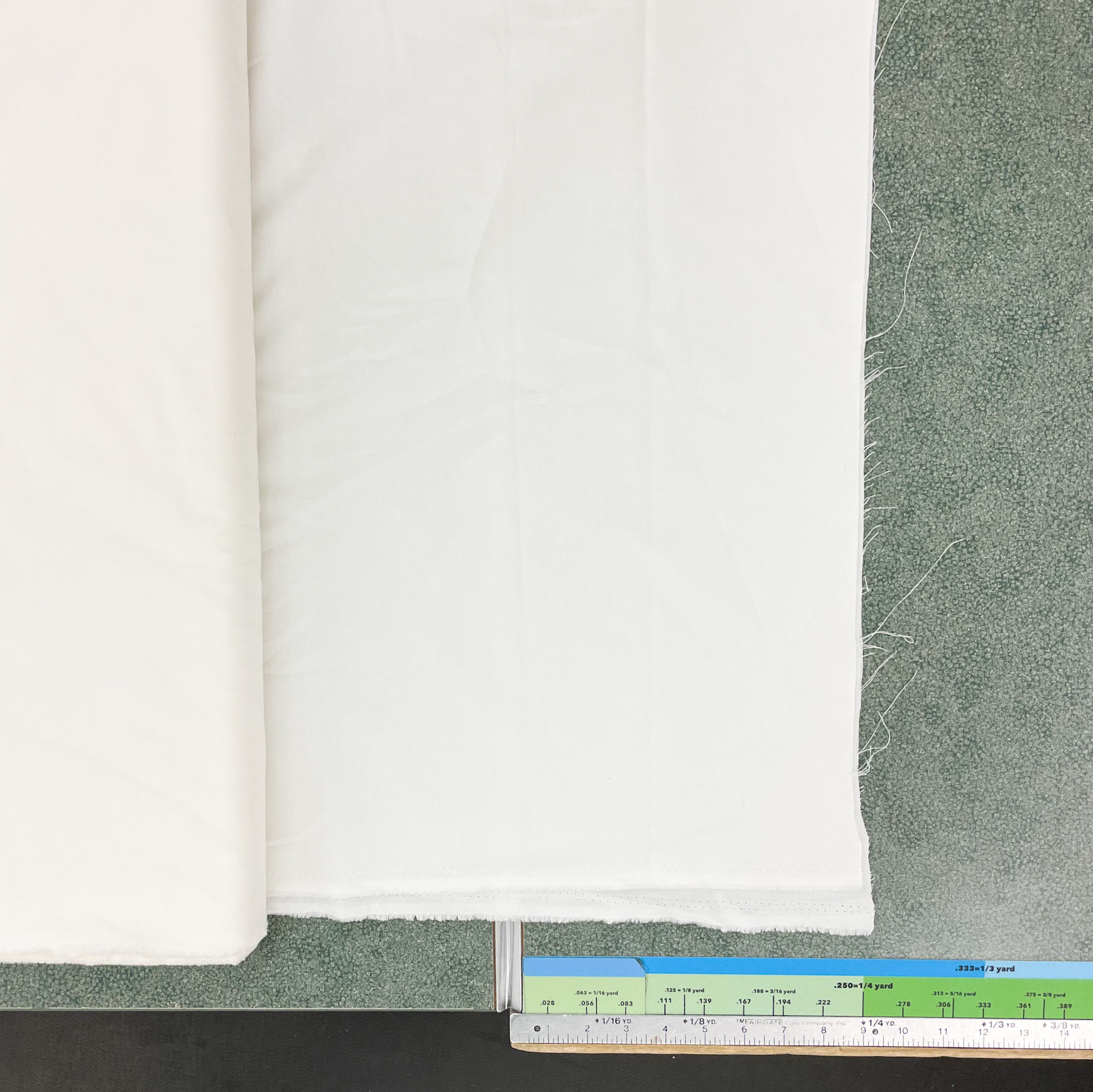 Image shows a cutting table with cream fabric positioned to be cut. At the bottom of the image is a ruler marking yard increments and inches. 