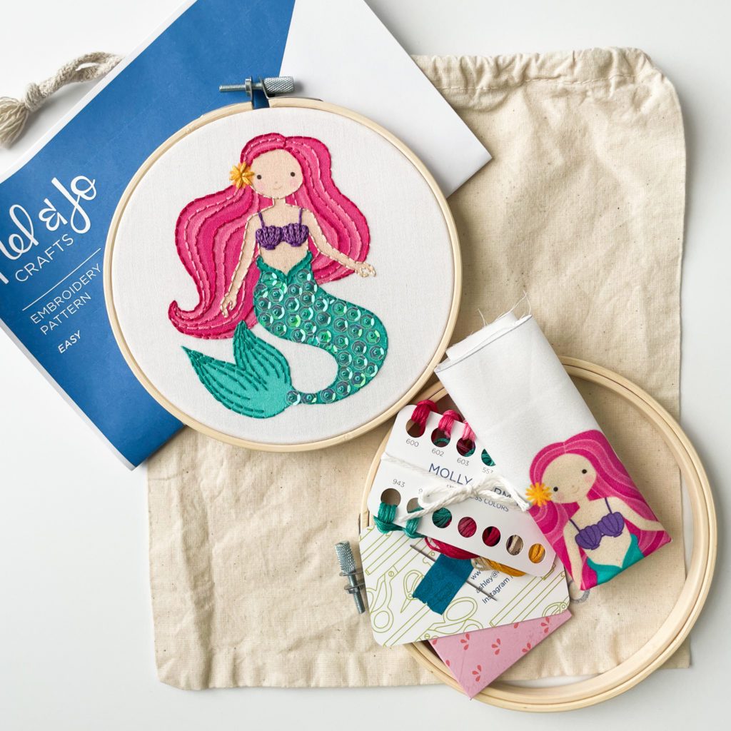 Image of an embroidery mermaid with pink hair. You see both a finished embroidery and the unfinished supplies 